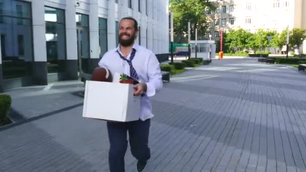Begrip ontslag van werk, aanwerving. Een werknemer loopt tussen kantoorgebouwen met een kartonnen doos en documenten met een bureau bloem. Mijn baan kwijtgeraakt door een fout. — Stockvideo