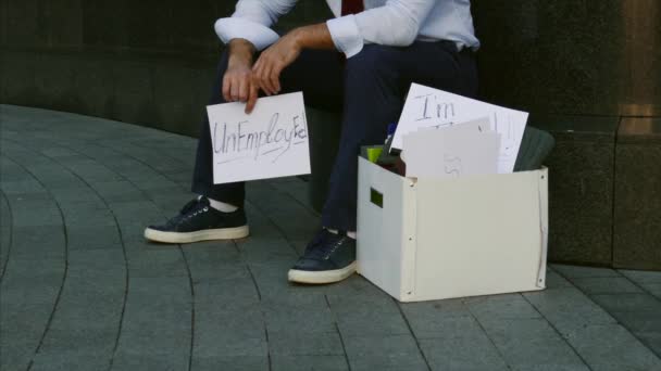 Werkloosheid, werkloze, ontslag, inkrimping. Een man zonder werk. Een jongeman zit op straat met een bord ontslagen, een baan nodig. — Stockvideo