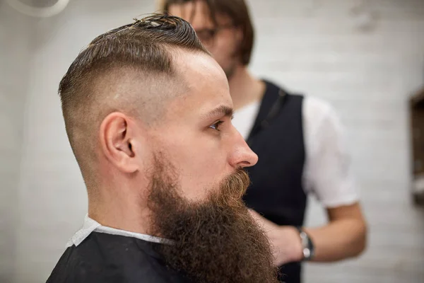 Um brutamontes na Barbearia moderna. Cabeleireiro faz penteado um homem com uma longa barba. Cabeleireiro mestre faz penteado com cortador de cabelo — Fotografia de Stock