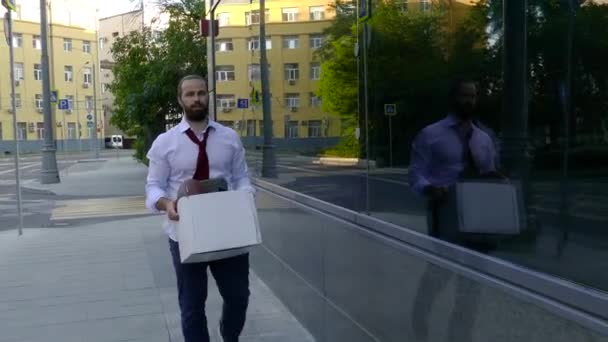 Un hombre guapo despedido con barba, un gerente con atuendo formal, molesto después de ser despedido, va junto con una caja de pertenencias personales. Primer plano, depresión, mal humor, mal día . — Vídeo de stock