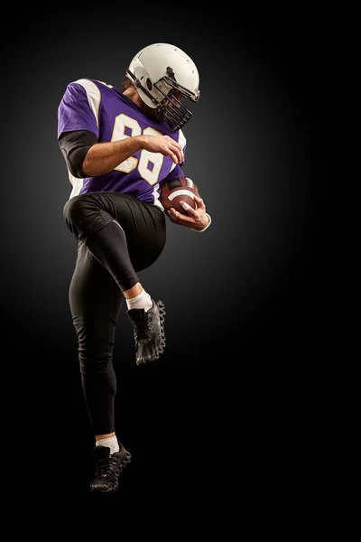 American football player holding a ball while jumping away from a strike. Black background, copy space. Violet with white sportswear.