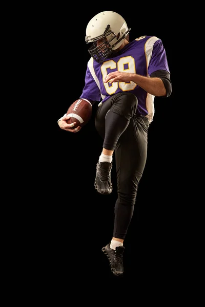 American football player holding a ball while jumping away from a strike. Black background, copy space. Violet with white sportswear.