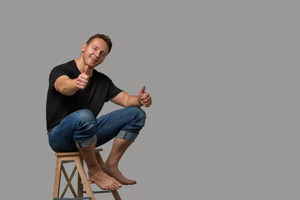 A man in a black T-shirt and jeans sitting on a chair shows with his hands on your text or your advertisement on a gray background. Concept banner for advertising, copy space, gray background — Stock Photo, Image