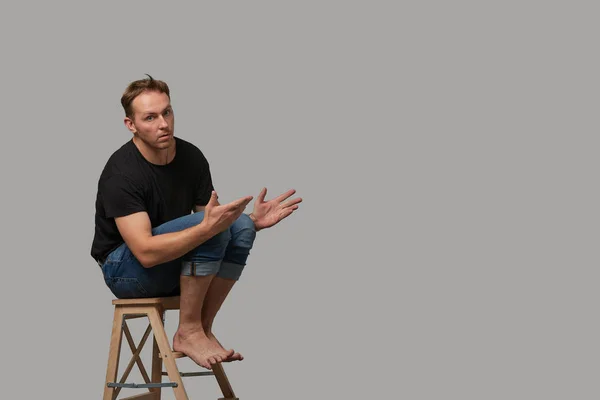 Een man in een zwart T-shirt en jeans zittend op een stoel toont met zijn handen op uw tekst of uw advertentie op een grijze achtergrond. Concept banner voor adverteren, kopieer ruimte, grijze achtergrond — Stockfoto