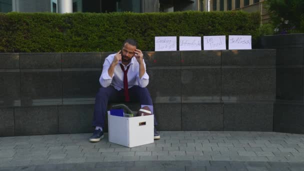 Ontslagen Business man zitten gefrustreerd en boos op de straat in de buurt van kantoorgebouw met doos van zijn bezittingen. Hij verloor het werk. Rond de gedecomponeerde kartons gelabeld, ontslagen, werkloos, werk nodig — Stockvideo