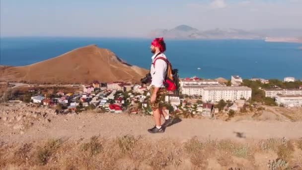 Hombre viajero con una mochila y una cámara, caminando por las montañas, con una gran vista de las montañas y el mar. Disparos desde arriba, el concepto de viaje, nuevas aperturas, libertad — Vídeos de Stock