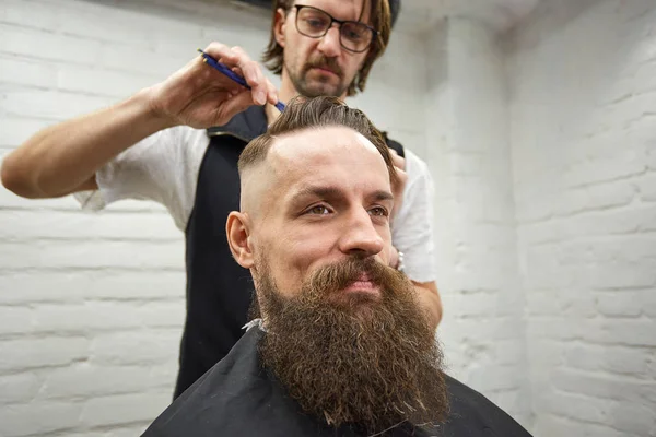 Master cuts hair and beard of men in the barbershop, hairdresser makes hairstyle for a young man