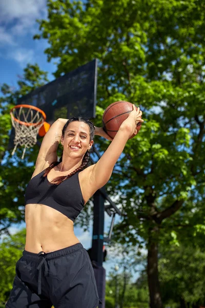 Hermosa chica latina deportiva con una pelota de baloncesto debajo del ring en una cancha de baloncesto callejera. Motivación deportiva, estilo de vida saludable, espacio para copiar, banner publicitario . — Foto de Stock
