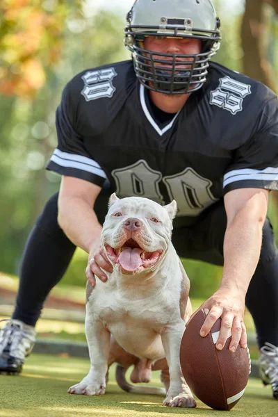 Jugador de fútbol americano con un perro posando en cámara en un parque. Copiar espacio, bandera deportiva. Concepto fútbol americano, deporte para la protección de los animales . — Foto de Stock