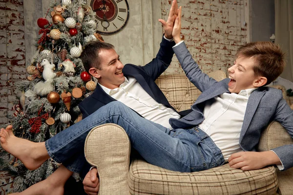Hijo y padre se regocijan con el telón de fondo de las decoraciones navideñas. El hijo y el padre están sentados en una silla y charlando alegremente en Nochebuena . — Foto de Stock