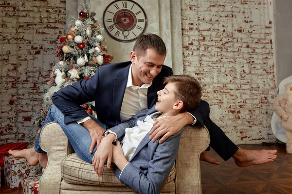 Hijo y padre se regocijan con el telón de fondo de las decoraciones navideñas. El hijo y el padre están sentados en una silla y charlando alegremente en Nochebuena . — Foto de Stock