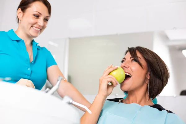 Портрет молодого стоматолога и ее пациента в кабинете стоматолога. Молодая дантистка держит зеленое яблоко в клинике . — стоковое фото