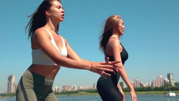 Dos mujeres haciendo los ejercicios deportivos a orillas del río en la ciudad. Los ejercicios al aire libre . — Vídeos de Stock