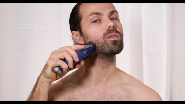 Hezký mladý muž s dlouhými vlasy si v koupelně holí vousy nůžkami. Krásné světlo, rozmazané pozadí. — Stock video