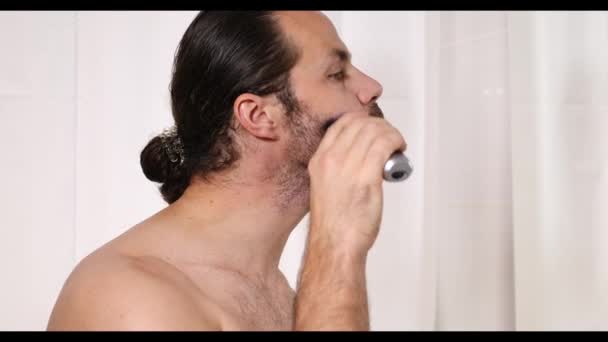 長髪のハンサムな若い男がバスルームでトリマーで髭を剃っています。美しい光、ぼやけた背景. — ストック動画