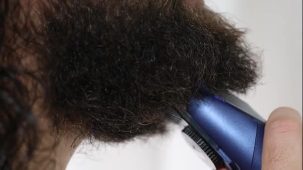 Handsome young man with long hair shaves his beard with a trimmer in the bathroom. Close-up. — Stock Video