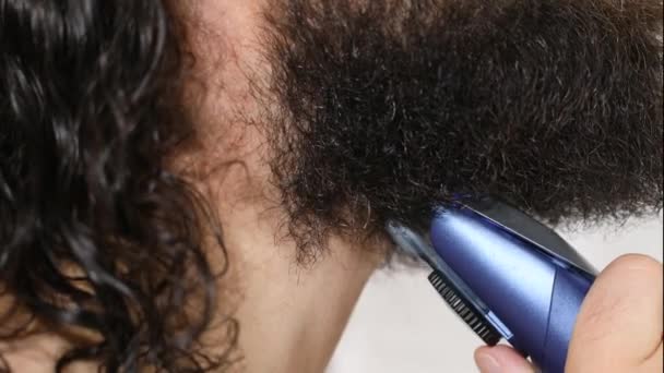 Handsome young man with long hair shaves his beard with a trimmer in the bathroom. Close-up. — Stock Video