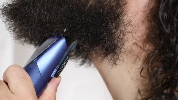 Handsome young man with long hair shaves his beard with a trimmer in the bathroom. Close-up. — Stock Video