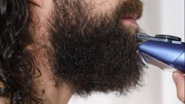 Handsome young man with long hair shaves his beard with a trimmer in the bathroom. Close-up. — Stock Video