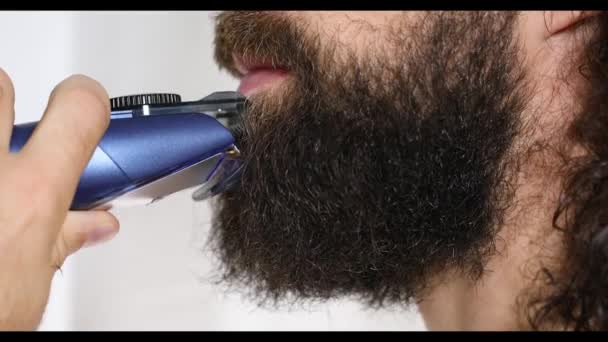 Knappe jongeman met lang haar scheert zijn baard met een trimmer in de badkamer. Close-up. — Stockvideo