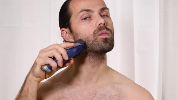 Hezký mladý muž s dlouhými vlasy si v koupelně holí vousy nůžkami. Krásné světlo, rozmazané pozadí. — Stock video