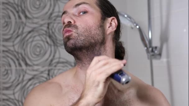 Knappe jongeman met lang haar scheert zijn baard met een trimmer in de badkamer. Mooi licht, wazige achtergrond. — Stockvideo