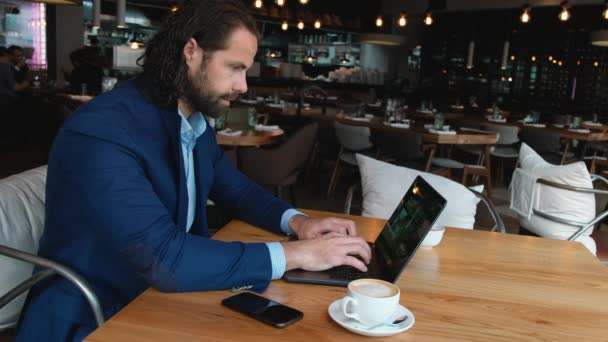 Joven hombre de negocios europeo sentado en una mesa en un café y escribiendo en un ordenador portátil mientras trabaja en un descanso para el café . — Vídeo de stock