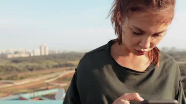 Una mujer joven y bonita se encuentra en una azotea en medio de una gran metrópoli durante una puesta de sol naranja en una noche de verano. Ella está chateando con estilo en un teléfono inteligente y mirando a la cámara . — Vídeos de Stock