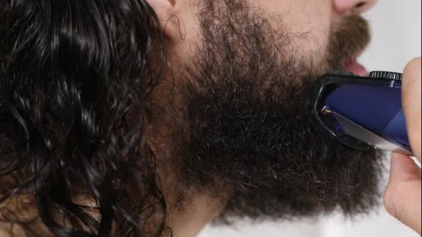 Handsome young man with long hair shaves his beard with a trimmer in the bathroom. Close-up. — Stock Video