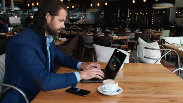 若いヨーロッパのビジネスマンがカフェのテーブルに座って、コーヒーブレイクで働いている間にノートパソコンを入力します。. — ストック動画