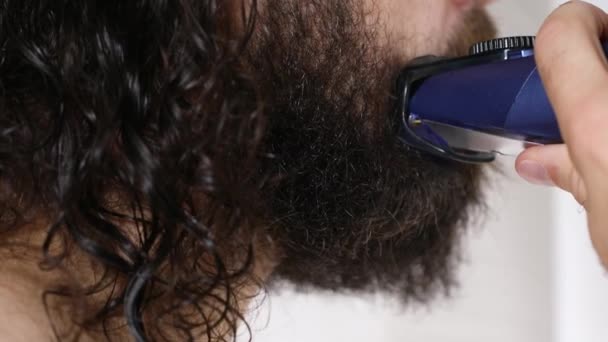 Handsome young man with long hair shaves his beard with a trimmer in the bathroom. Close-up. — Stock Video