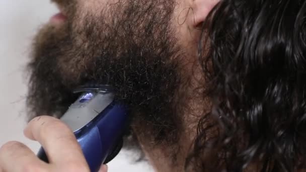 Handsome young man with long hair shaves his beard with a trimmer in the bathroom. Close-up. — ストック動画