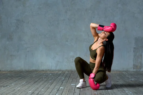 Modelo menina bonita posando na câmera em luvas de boxe em luz rosa. Menina sentada no chão em um fundo de concreto cinza. Banner de esportes, espaço de cópia . — Fotografia de Stock