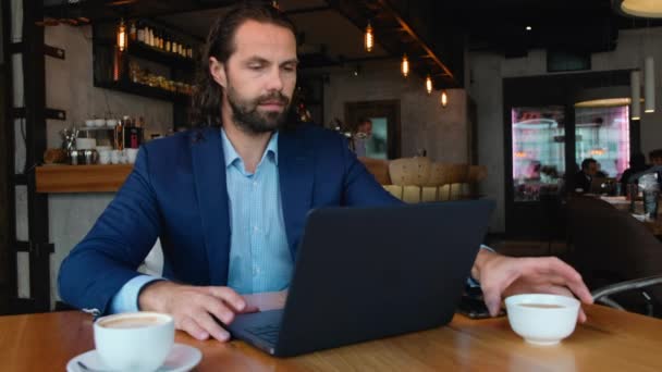 Fiatal európai üzletember ül egy asztalnál egy kávézóban, és gépel egy laptopon, miközben a kávészüneten dolgozik.. — Stock videók