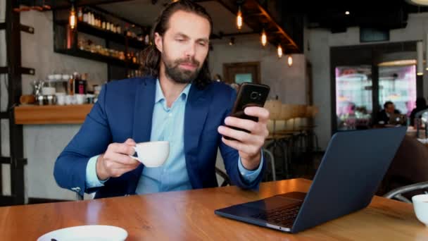 En ung affärsman som sitter vid ett bord på ett kafé och använder telefonen. Framgångsrika människor, dagliga rutiner. Underhållande, avkopplande, pratande. — Stockvideo