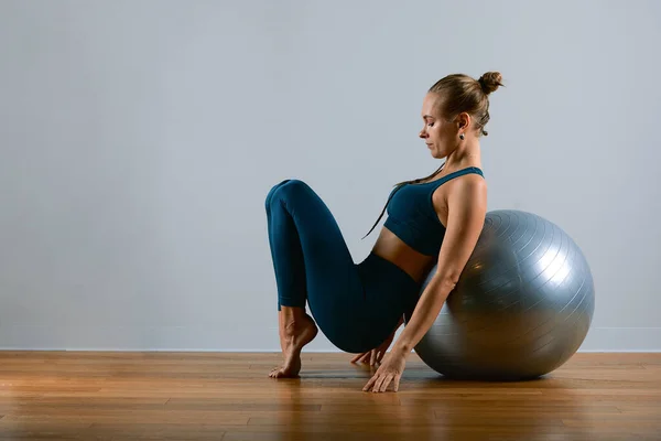 Young, beautiful, athletic girl doing exercises on fitball in the gym on a gray background. Sporting Slavic girl in a blue, green suit. Copy space, gray background, sport banner for advertising.