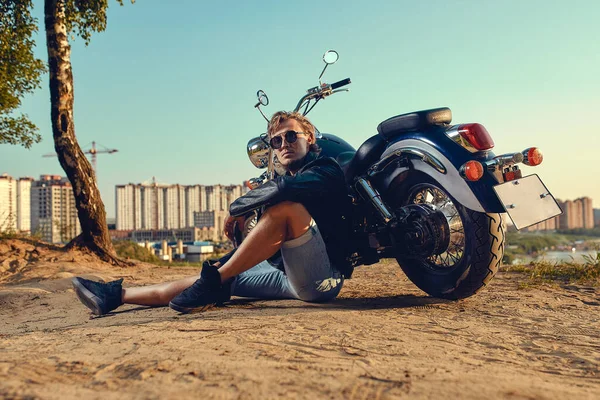 Sexig biker man bär jeans och läder jacka sitter avslappnad av sin motorcykel — Stockfoto