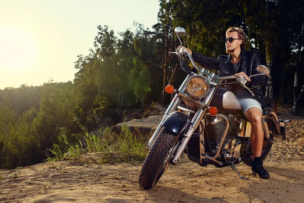 Brutal ung man i solglasögon, blå jeans och en svart skinnjacka sitter på den egna motorcykeln — Stockfoto