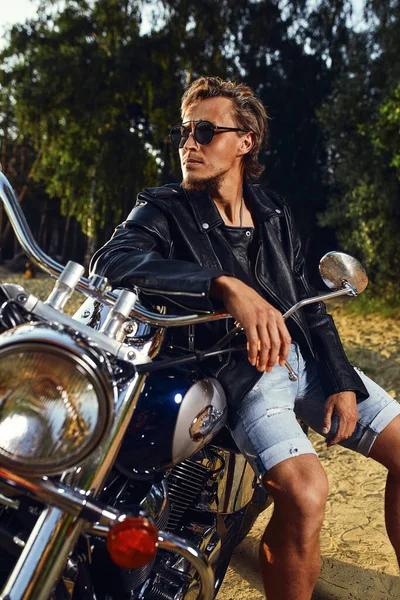 Brutal young man in sunglasses, blue jeans and a black leather jacket sitting on the custom motorcycle