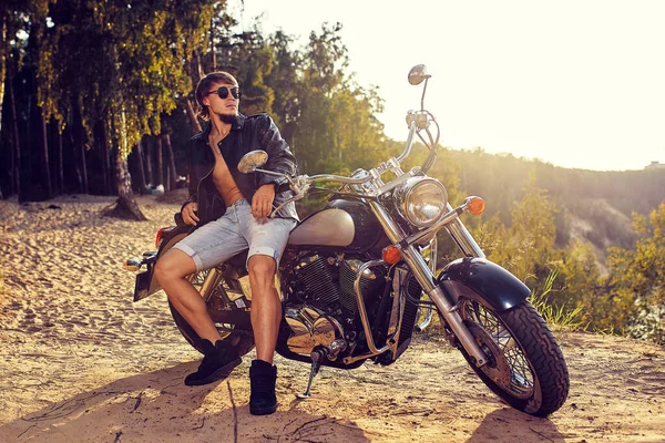 Joven brutal en gafas de sol, vaqueros azules y una chaqueta de cuero negro sentado en la motocicleta personalizada — Foto de Stock