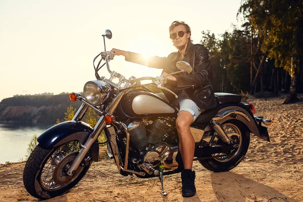Brutal ung man i solglasögon, blå jeans och en svart skinnjacka sitter på den egna motorcykeln — Stockfoto
