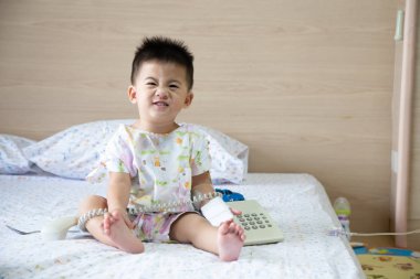 Asian baby boy smiling and wear patients suits sitting on bed in the room at child department in the hospital. Children with infectious diseases IPD, Invasive Pneumococcal Disease and under 2 years clipart