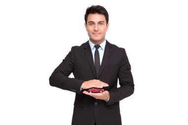 Portrait of a charming businessman dressed in suit holding toy car while standing isolated over white background, Car insurance and car services concept clipart