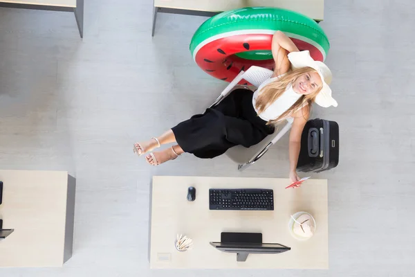 Vista superior de la mujer de negocios sentada en el escritorio durante el trabajo con el ordenador en la oficina y preparar bolsas de viaje y anillos de goma para los viajes al mar, plan de vacaciones de verano y el concepto de vacaciones largas — Foto de Stock