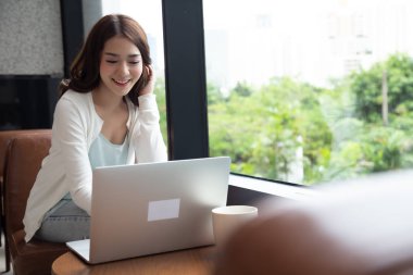 Güzel gülümseyen Asyalı kadın blogger rahat oturan giyinmiş ve kafe ve restoranda ahşap masada dizüstü bilgisayar kullanarak