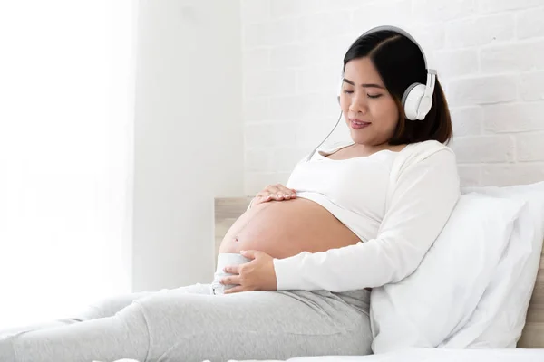 젊은 아시아 임신 한 여자 듣기 음악 과 앉아 에 침대 에 이 침실 — 스톡 사진