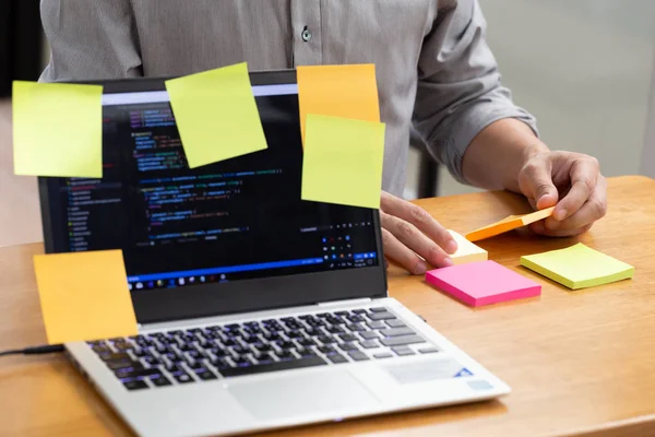 Tasks on sticky note cards on laptop computer with source code program of business software development planning and programmer holding short note paper on hand