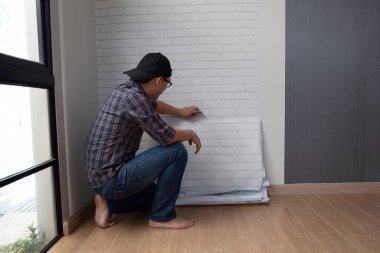 Genç Asyalı adam evdeki duvara duvar kağıtlarını yapıştırıyor. Kendini yenileme konsepti.