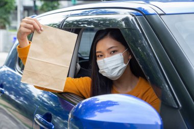Asian woman wearing mask and holding paper bag of fast food through the window car. Drive thru food service concept clipart