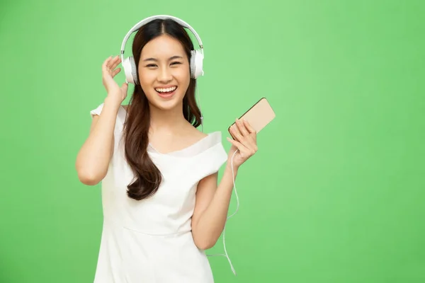 若いアジアの美しさの女性は緑の背景に隔離されたスマートフォン上のプレイリストの歌アプリケーションでヘッドフォンで踊り 音楽を聴く — ストック写真